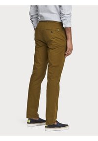 Pantalon en coton marron avec une coupe slim, doté d'une poche arrière avec bouton et d'un étiquette logo verte. Porté avec des mocassins navy.