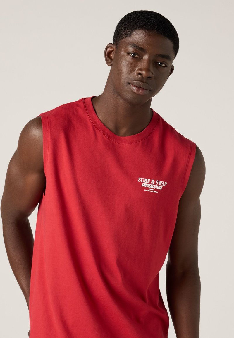 Junger Mann mit kurzen Haaren trägt ein rotes ärmelloses Shirt mit weißem Schriftzug "SURF & SWAP INVITATIONAL" auf der Brust, neutraler Hintergrund.