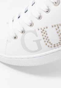 Chaussure de sport blanche en matériau synthétique, avec des clous ronds en argent et un logo gris clair. Semelle en caoutchouc texturé avec un design à lacets.