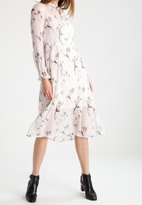 Floral-print, long-sleeve dress in light pink with a flowy silhouette; button-down front and textured fabric. Worn with black ankle boots.