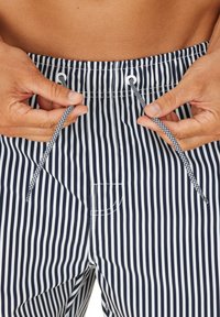 Navy- und weiß gestreifte Badehosen mit einem Kordelzugbund, ausgestattet mit einer kleinen Tasche und glatter Textur. Hände passen den Kordelzug an.