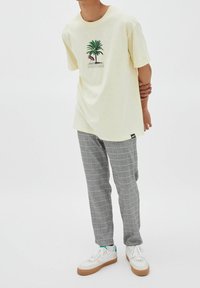 T-shirt di cotone gialla con grafica di palma e fenicottero, abbinata a pantaloni a quadri grigi e sneakers bianche con suole marroni.