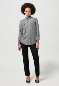 Camisa de mezclilla gris con botón en la parte delantera y dos bolsillos en el pecho, combinada con pantalones negros de pierna recta y sandalias de tacón negras.