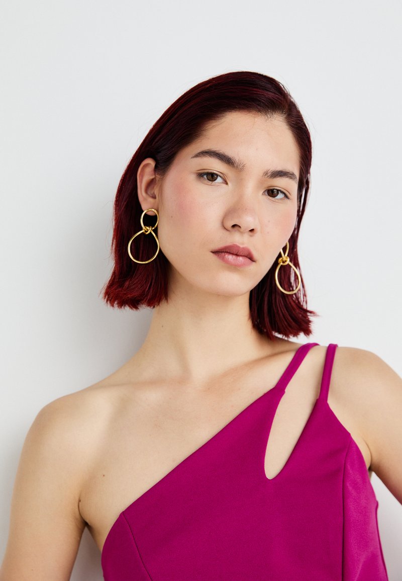 Vestido fucsia sin mangas con un corte asimétrico y un corte lateral. Pendientes de aro circulares dorados complementan el look. Textura suave.
