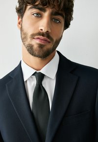 Giovane uomo con capelli ricci e barba, indossa una camicia bianca, cravatta nera e blazer blu scuro su uno sfondo bianco uniforme.
