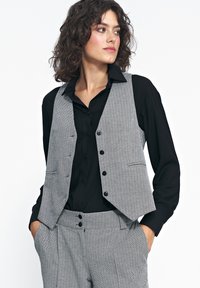 Czarna kamizelka w szaro-czarną kratę houndstooth z zaokrąglonym dołem, pięcioma guzikami i bocznymi kieszeniami, noszona na czarnej koszuli. Model ma kręcone włosy.