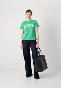 Zelené bavlněné tričko s bílým textem "BARBOUR", spárované s tmavými denimovými rozšířenými džíny a černou koženou taškou přes rameno. Jednoduchý, každodenní outfit.
