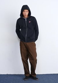 Juoda zip-up gobtuvu su rožine logotipu, derinama su rudos spalvos kelnėmis. Gobtuvas turi raištelių ir šonines kišenes. Juodi batai užbaigia įvaizdį.