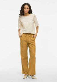 Top de encaje crema con mangas cortas y festoneadas; pantalones de pierna ancha mostaza; zapatos de tacón altos beige; modelo de pie con las manos en los bolsillos.