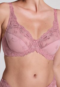 Svetlo roza modrček s cvetličnim čipkastim detajlom, mrežastimi skodelicami in valovitimi robovi. Prilagodljive naramnice in pentlja na sredini.