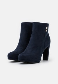 Botas de tobillo de ante sintético azul marino con punta redondeada, tacón alto en bloque y dos detalles decorativos de botones dorados en el lado.