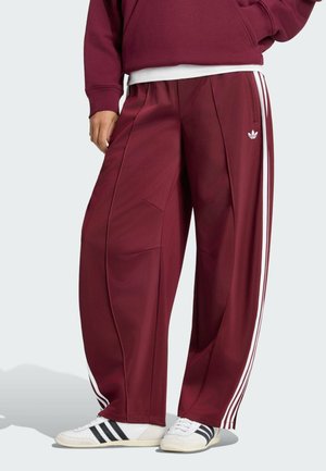 Persona con pantalones deportivos Adidas granate con franjas blancas a los lados y zapatillas blancas con franjas negras.