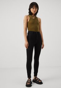 Jauna moteris su juodais plaukais, vilkinti alyvuogių spalvos be rankovių marškinėlius su išpjova, juodas siauras kelnes, juodas šlepetes ir mėlyną akių makiažą, stovinti.