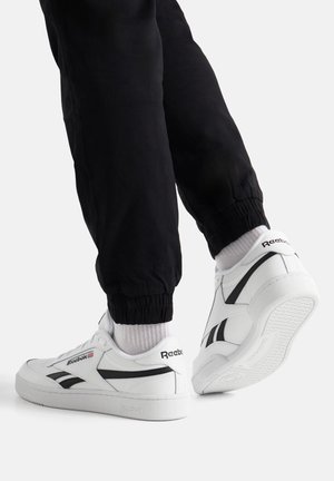 Osoba nosící bílé tenisky Reebok s černými detaily, bílé ponožky a černé teplákové kalhoty na jednobarevném pozadí.