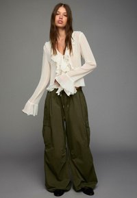 Bílá blůza s volánkovým výstřihem a manžetami, ve spojení se zelenými cargo kalhotami s velkými bočními kapsami. Černé boty dokončují celý outfit.