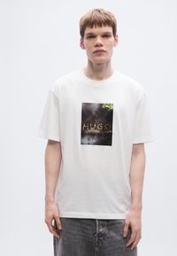 Άσπρο T-shirt με κ οντά μανίκια που διαθέτει ένα μαύρο τετράγωνο σχέδιο με τη λέξη "HUGO" σε καφέ χρώμα, περιτριγυρισμένο από πράσινα φύλλα και ένα υφασμάτινο φόντο.