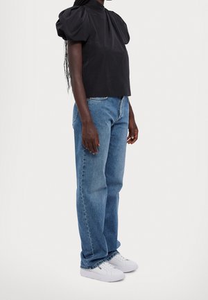 Femme debout portant un haut noir à manches bouffantes, un jean droit bleu et des baskets blanches devant un fond uni.