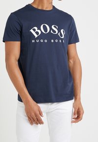 Laivansinis lygių rankovių marškinėliai, pagaminti iš medvilnės, su užrašu "BOSS HUGO BOSS" balta spalva per krūtinę; standartinio apvalaus kaklo dizainas.