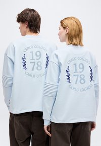 Δύο άτομα φορούν ανοιχτό μπλε φούτερ με την επιγραφή "CARLO COLUCCI 1978" και σχέδια δάφνης στην πλάτη, συνδυασμένα με σκούρα παντελόνια.