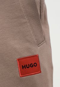 Καφέ ύφασμα με πλαϊνή τσέπη και ένα κόκκινο ορθογώνιο σημείο με την ετικέτα «HUGO» ραμμένο κοντά στη ραφή της τσέπης.