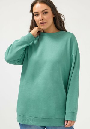 Kvinde med langt brunt hår iført oversized teal sweatshirt og blå jeans, poserende med hånden nær halsen mod en hvid baggrund.