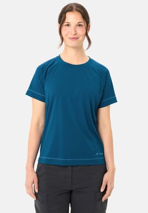 Donna in piedi, che indossa una maglietta atletica a maniche corte color teal e pantaloni cargo scuri, con i capelli raccolti e un leggero sorriso.