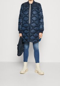 Tmavě modrá péřovka s prošívaným vzorem, žebrovanými manžetami a předním zipem. Doplňuje ji pruhovaný top, modré džíny a krémové boty.