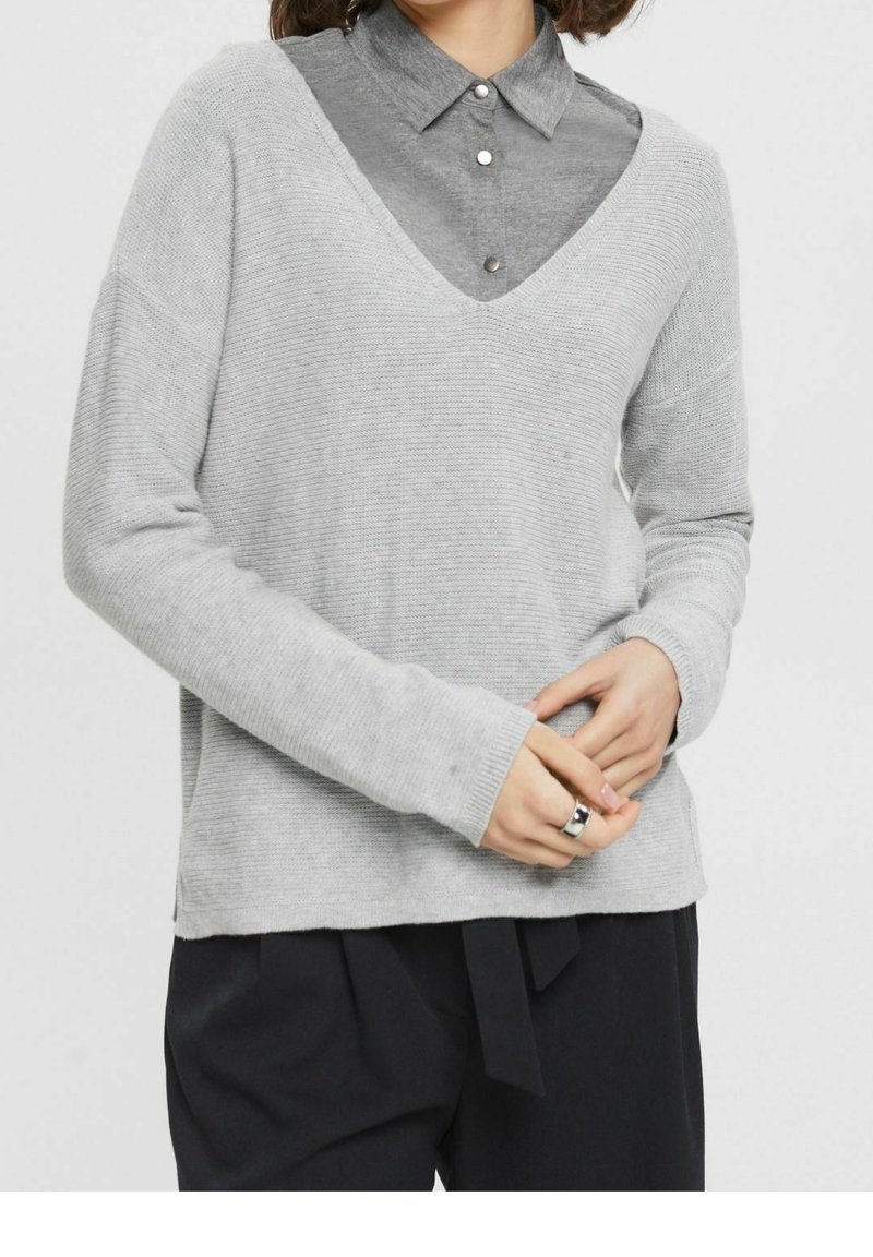 Femme portant un pull gris clair à col en V sur une chemise grise boutonnée, avec un pantalon noir, joignant les mains avec une bague en argent à la main droite.