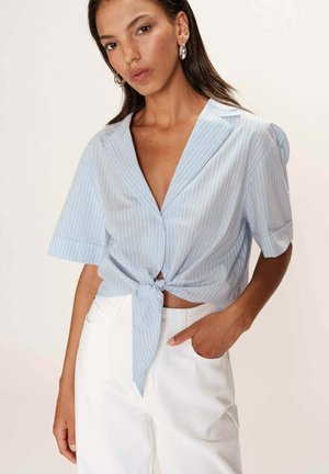 Chemise à manches courtes rayée bleue et blanche avec un nœud à la taille, dotée d'un décolleté en V profond et d'une texture de tissu douce. Associée à un pantalon blanc.