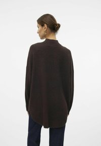 Pull oversized marron foncé avec un col montant, des épaules tombantes et un ourlet arrondi. Texture douce et coupe décontractée. Porté avec un jean bleu.