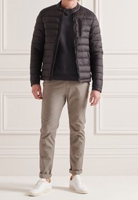 Veste matelassée noire, fermeture éclair à l'avant, col montant et coupe ajustée. Associée à un pantalon beige clair et des baskets blanches, sur un fond neutre.