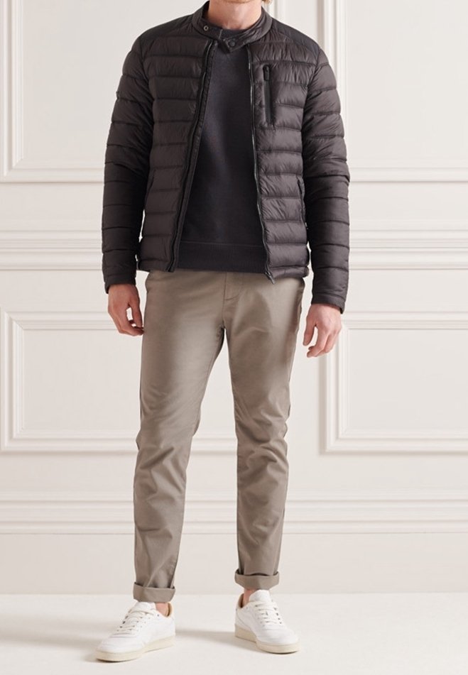 Veste matelassée noire, fermeture éclair à l'avant, col montant et coupe ajustée. Associée à un pantalon beige clair et des baskets blanches, sur un fond neutre.
