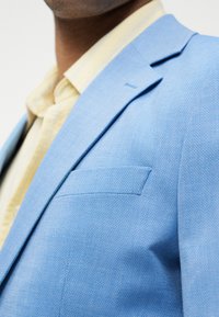 Άνδρας που φορά ανοιχτό γαλάζιο σακάκι πάνω από ένα απαλό κίτρινο πουκάμισο με γιακά, εμφανιζόμενος από το στήθος και πάνω, μπροστά σε απλό λευκό φόντο.