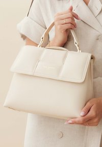 Sac à main en cuir beige au design structuré, avec une anse supérieure et un logo doré. Il comporte un système de fermeture à rabat et une surface texturée.