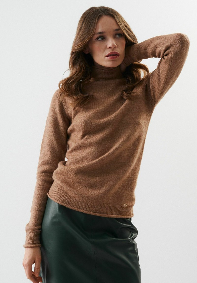 Pull à col roulé marron en tissu tricoté doux ; design ajusté avec des poignets et un ourlet côtelés ; porté avec une jupe en cuir vert foncé.