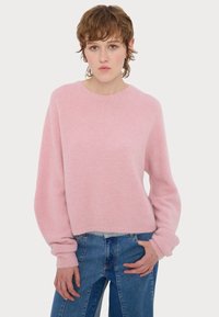 Νεαρό άτομο με κοντά καστανά μαλλιά που φοράει ένα φαρδύ ροζ πουλόβερ και δίχρωμο τζιν μπλε, στέκεται �μπροστά σε λευκό φόντο.