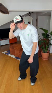 Fehér póló zöld logóval, fekete nadrág és fekete-fehér sapka. Ezüst cipők és texturált barna kanapé látható a háttérben.