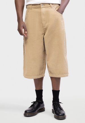 Egy személy, aki bézs színű, térdig érő kordnadrágot, fekete zoknit és fekete bőr fűzős cipőt visel, egyik kezét zsebében tartva áll.