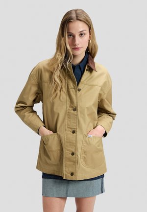 Jeune femme aux longs cheveux blonds portant une veste beige boutonnée sur une chemise sombre et une courte jupe en jean bleue, les mains dans les poches de la veste.