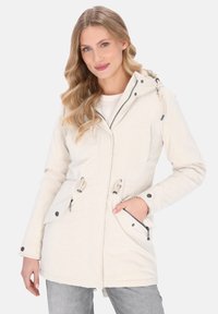 Kvindelig med langt bølget blond hår iført en lys beige hættetrøje med lynlås og knapdetaljer, poserende med den ene hånd i lommen.