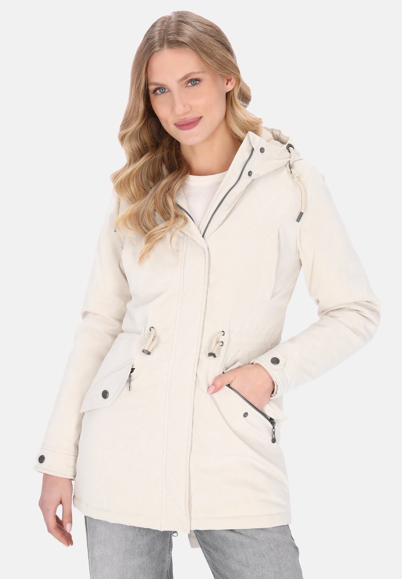 Kvindelig med langt bølget blond hår iført en lys beige hættetrøje med lynlås og knapdetaljer, poserende med den ene hånd i lommen.