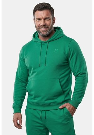 Uomo con barba che indossa una felpa verde con cappuccio e pantaloni della tuta abbinati, sorridente, con una mano in tasca su uno sfondo semplice.
