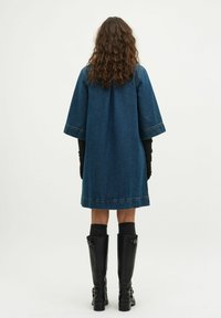 Jeanskleid mit lockerer Passform, kurzen Ärmeln und einer dunkelblauen Wäsche. Getragen mit hohen schwarzen Stiefeln und knielangen Socken. Rückansicht.
