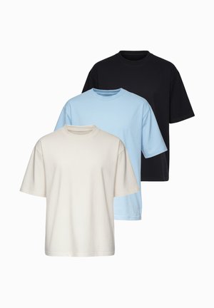 Homme portant un t-shirt beige oversize, un jean bleu clair, des baskets blanches et une montre-bracelet, debout devant un fond clair uni.