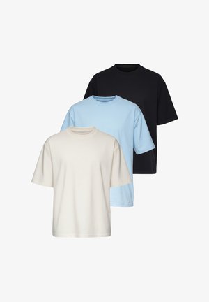 Man draagt beige oversized t-shirt, lichtblauwe jeans, witte sneakers en een polshorloge, staand tegen een neutrale lichte achtergrond.