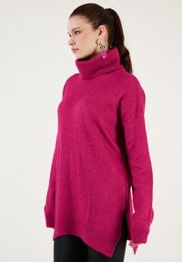 Magenta oversize poolo-kaulusneule, jossa on ribbbatut hihansuut ja helma, sivuliukkuja lisämukavuuden vuoksi sekä pehmeä, teksturoitu pinta.
