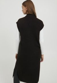 Gilet noir en maille côtelée, sans manches et long, avec un col montant et des fentes sur les côtés, porté sur une chemise blanche à manches longues. Texture lisse, silhouette ajustée.