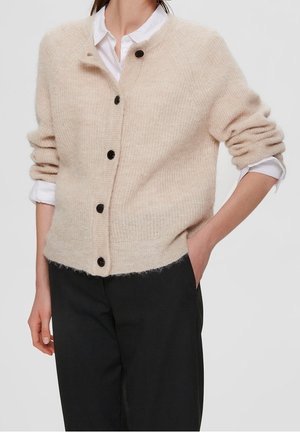 Beige gebreide cardigan, fluffy textuur, ronde hals, zes donkere knopen, geribbelde boorden, iets losse pasvorm, gedragen over een wit shirt.