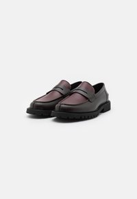 Mocassins en cuir bordeaux avec des semelles en caoutchouc noires contrastantes, présentant un design cousu et une lanière sur le dessus pour une touche de détail supplémentaire.