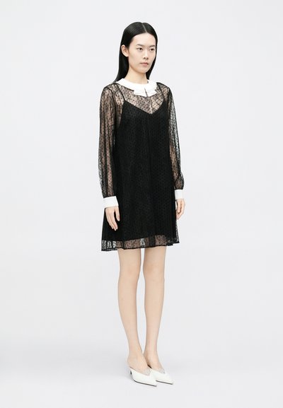 Svart blondekjole med lange, gjennomsiktige ermer og hvit krage. Glatt tekstur og knelengde silhuett. Sikker passform ved kanten.